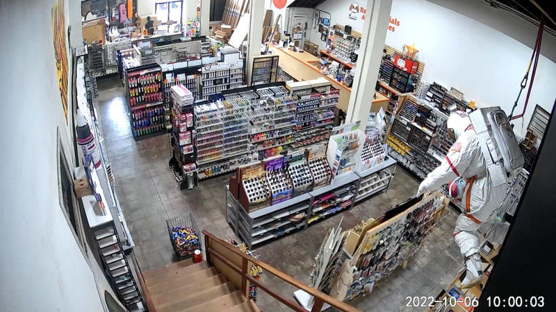 View of an art supply store from a security camera. Aisles of shelving and store displays are visible, a sculpture of an astronaut is hanging from the ceiling near the foreground.