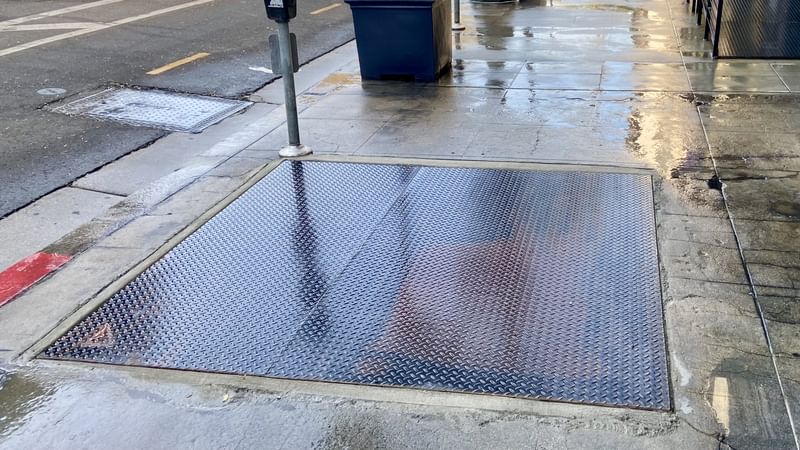 An access hatch embedded on the sidewalk on Main St that has been newly repaired. The ground is wet.